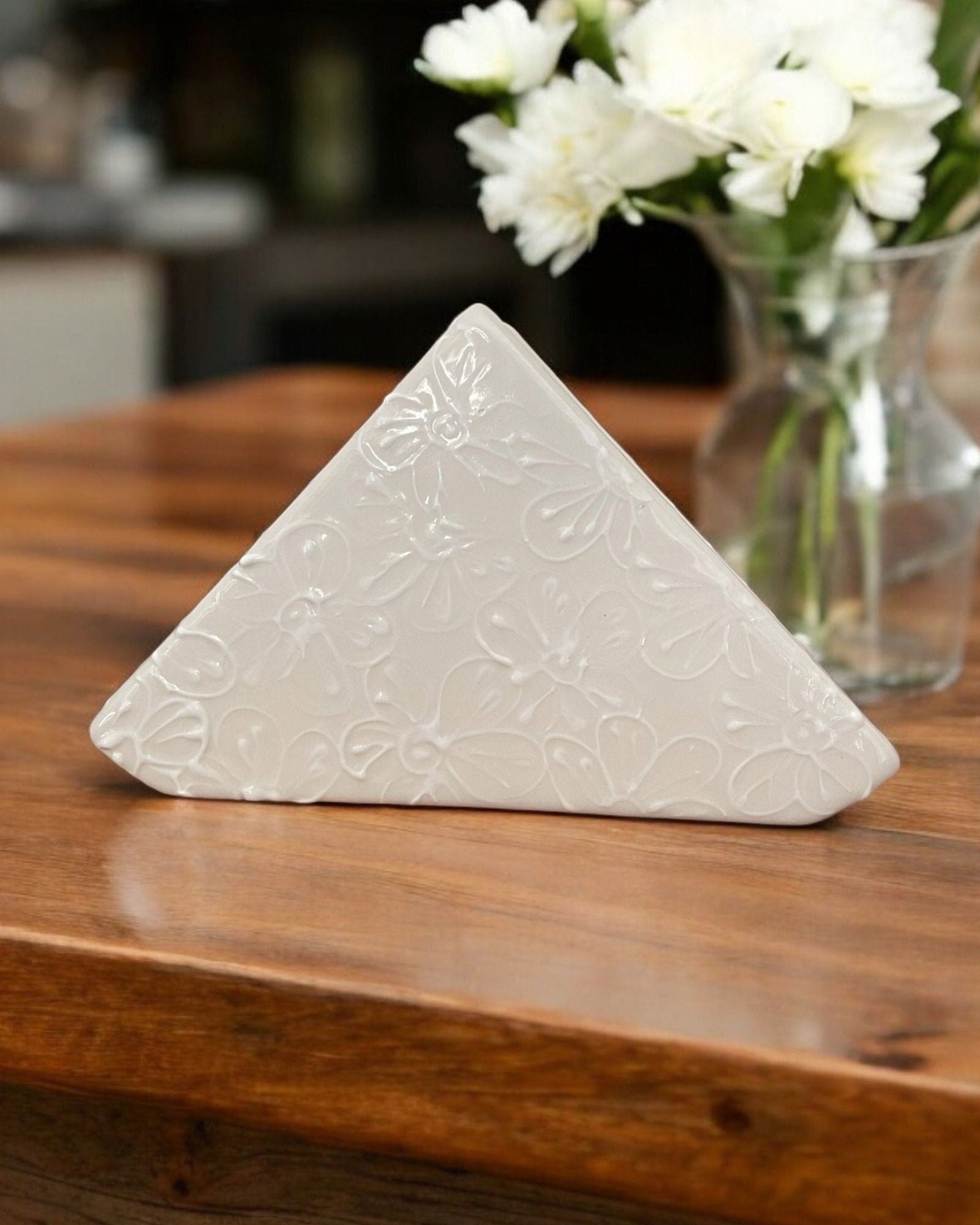 A triangular white ceramic napkin holder with painted floral patterns displayed on a wooden table.