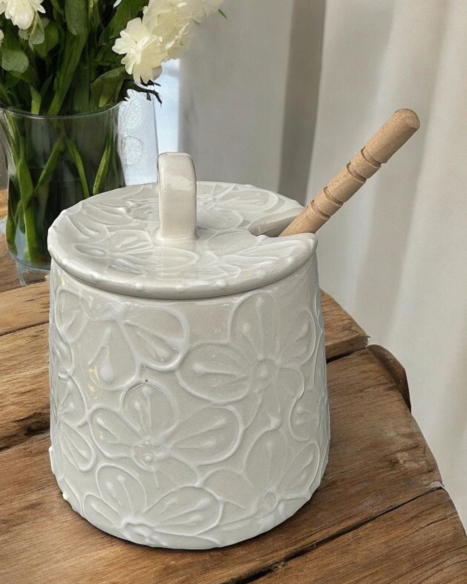 A cream-colored  ceramic honey pot jar with a lid and a wooden spoon, decorated with white floral motifs, set on a rustic wooden table with blurred purple and blue hydrangeas in the background.