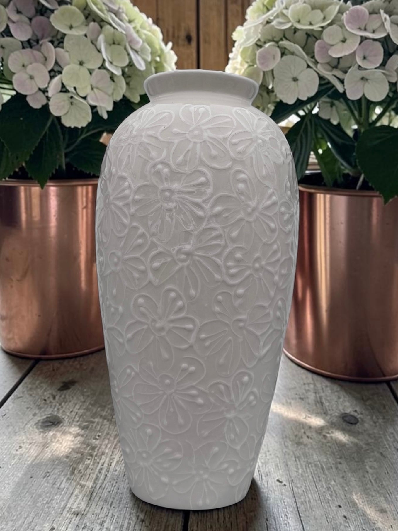 A white ceramic vase with an embossed floral pattern, standing on a rustic wooden surface with copper planters and white hydrangeas in the background.