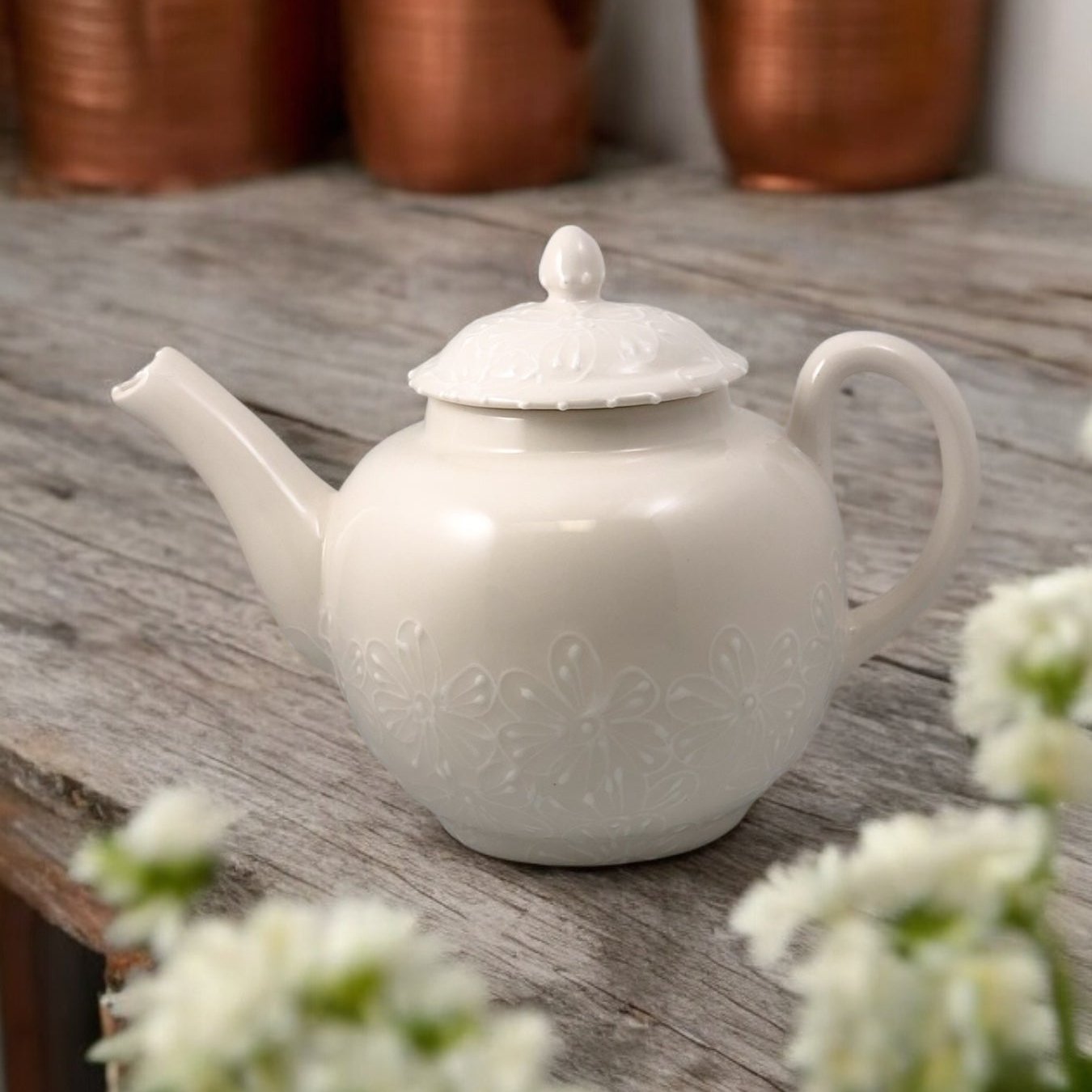 A white ceramic teapot with hand-painted floral designs, displayed on a wooden surface with a bouquet of white flowers beside it.