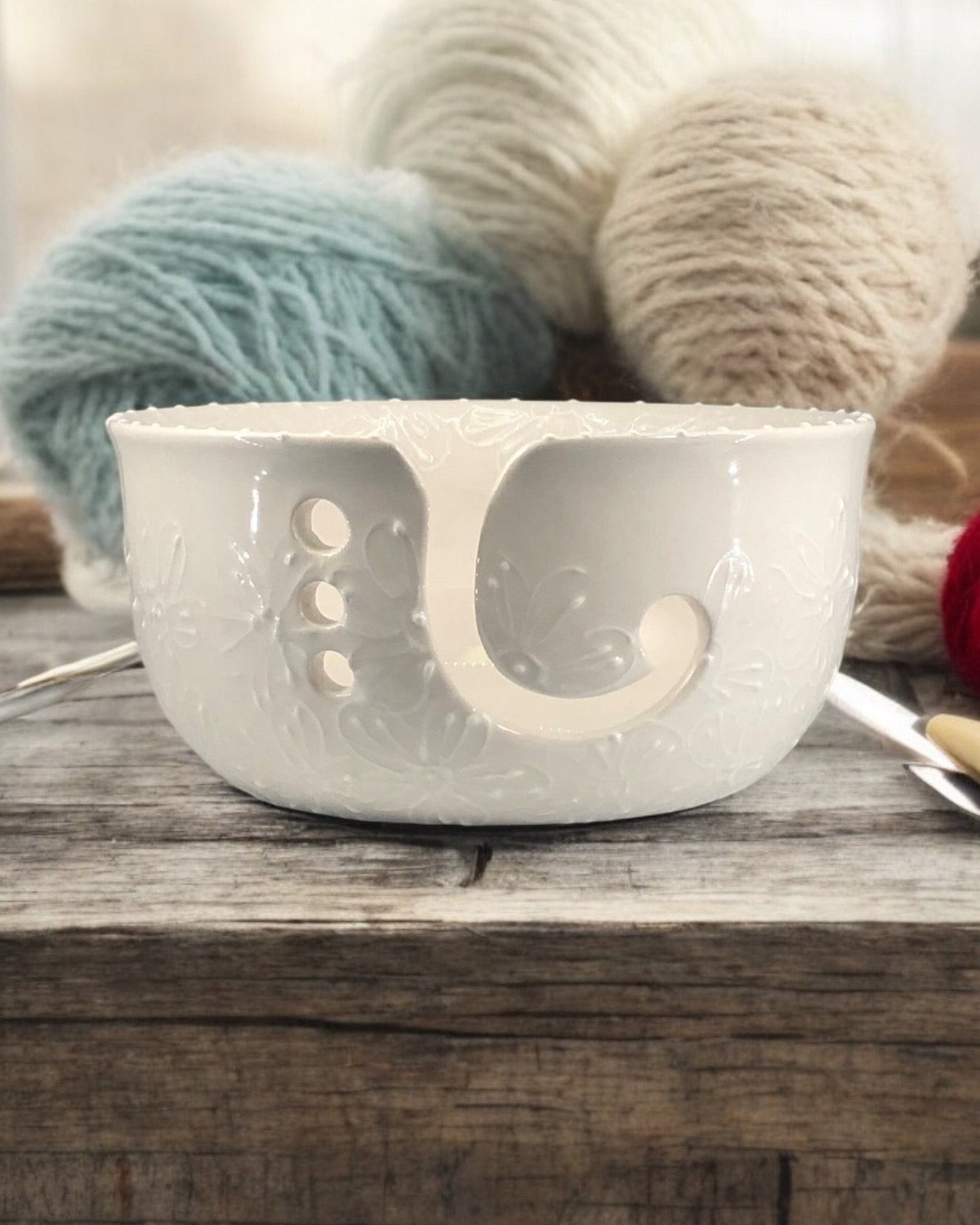 A hand-painted ceramic knitting bowl with white designs, containing yarn balls and knitting tools, placed on a wooden surface. Ladybird Ceramics Canada