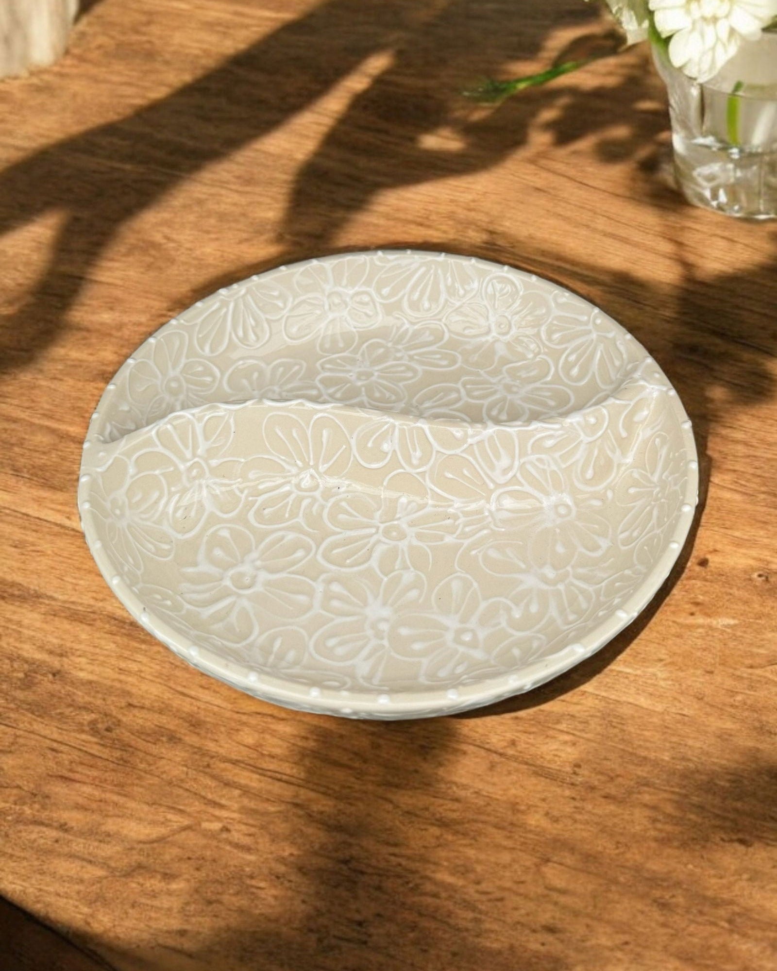 A ceramic two-compartment serving Bowl with a white base decorated with a white floral pattern, set on a white wooden surface with potted flowers in the background.