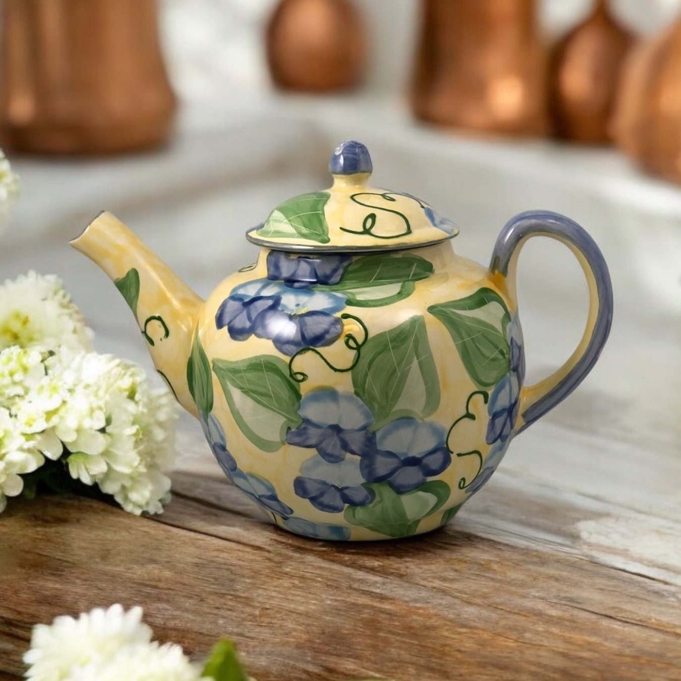 A white ceramic teapot with hand-painted floral designs, displayed on a wooden surface with a bouquet of white flowers beside it.