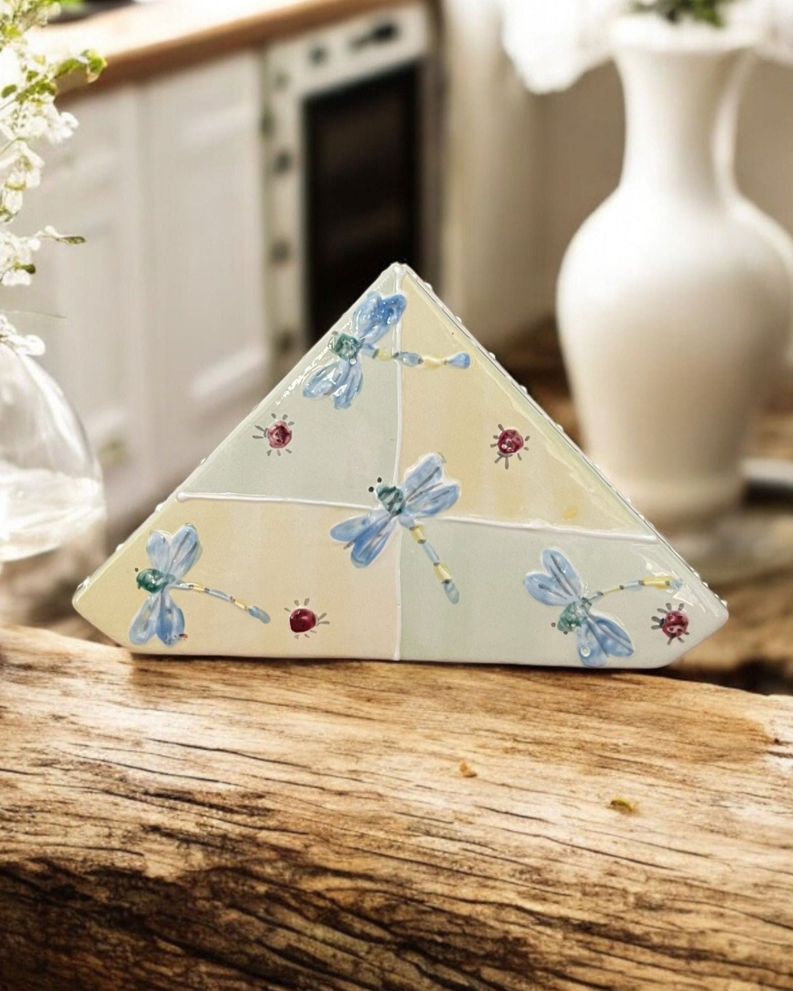 A triangular white ceramic napkin holder with painted floral patterns displayed on a wooden table.