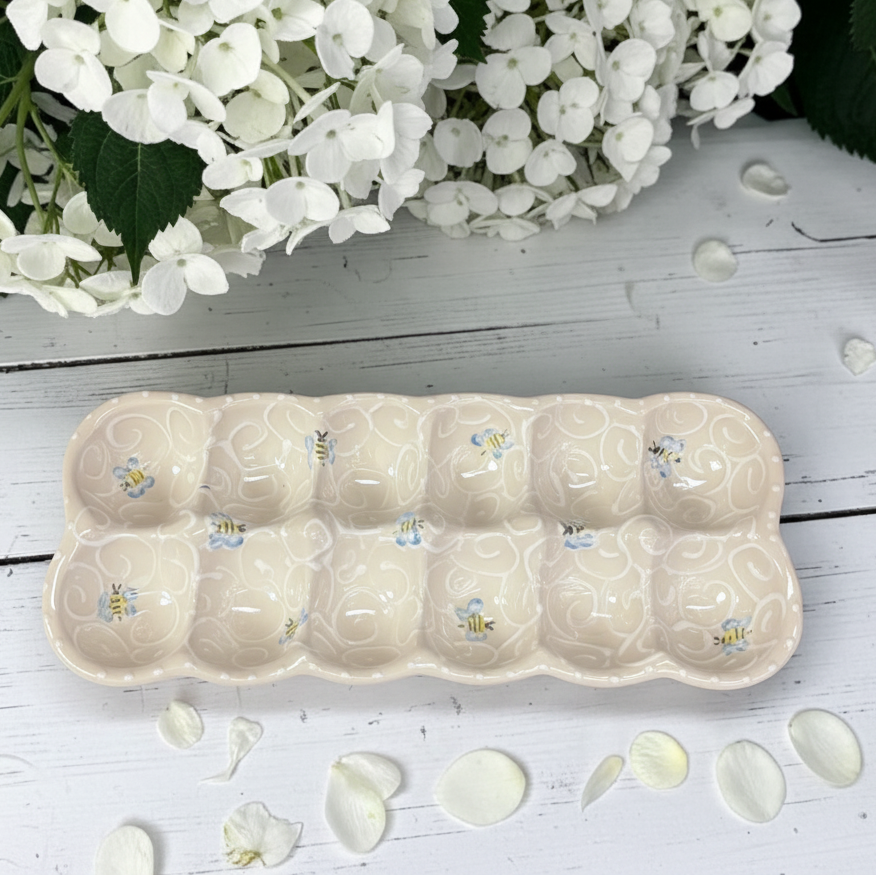 A ceramic 8-compartment egg tray in a warm cream color with a subtle swirl pattern and small bumblebee motifs, styled on a white wooden surface with white flowers and petals.