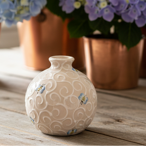 A small round ceramic vase with a textured embossed swirl pattern and small painted bees, set on a rustic wooden table with blurred blue and purple hydrangeas in copper pots behind it.