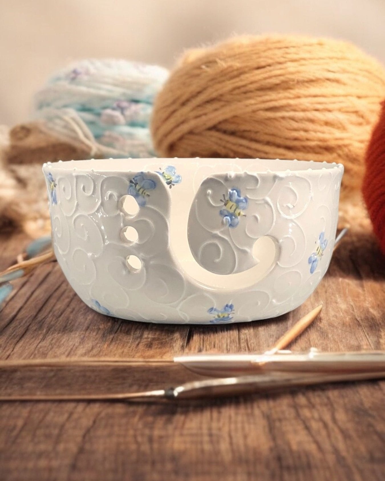 A hand-painted ceramic knitting bowl with blue bee painted design, containing yarn balls and knitting tools, placed on a wooden surface. Ladybird Ceramics Canada