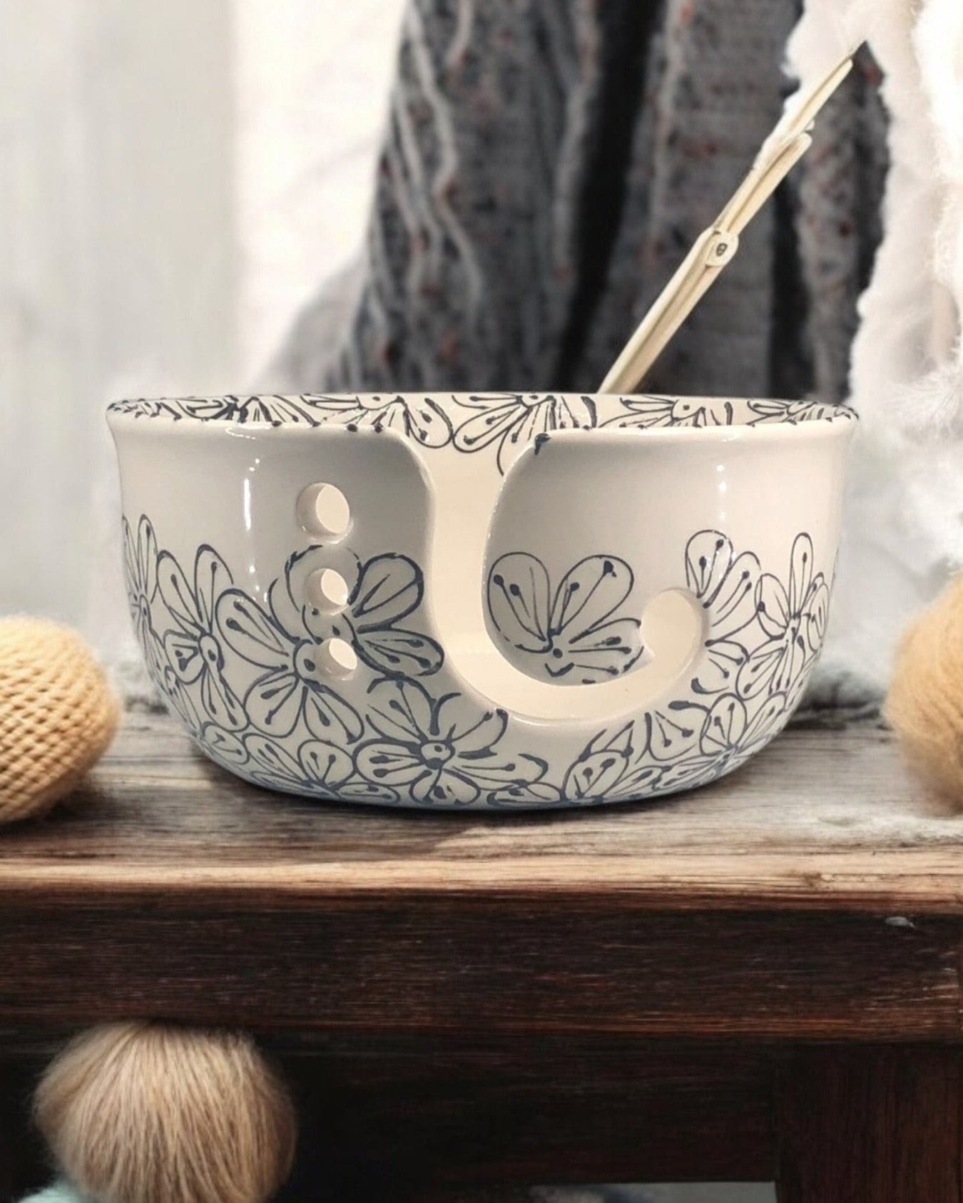 A hand-painted ceramic knitting bowl with black and white floral painted design, containing yarn balls and knitting tools, placed on a wooden surface. Ladybird Ceramics Canada