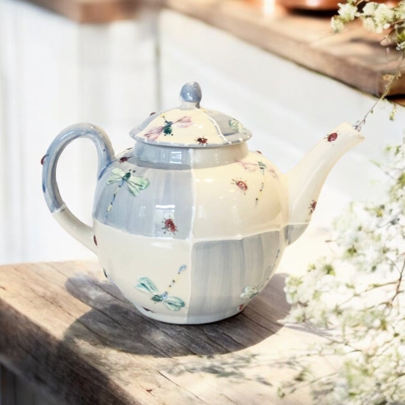 A white ceramic teapot with hand-painted floral  and dragonfly designs, displayed on a wooden surface with a bouquet of white flowers beside it.