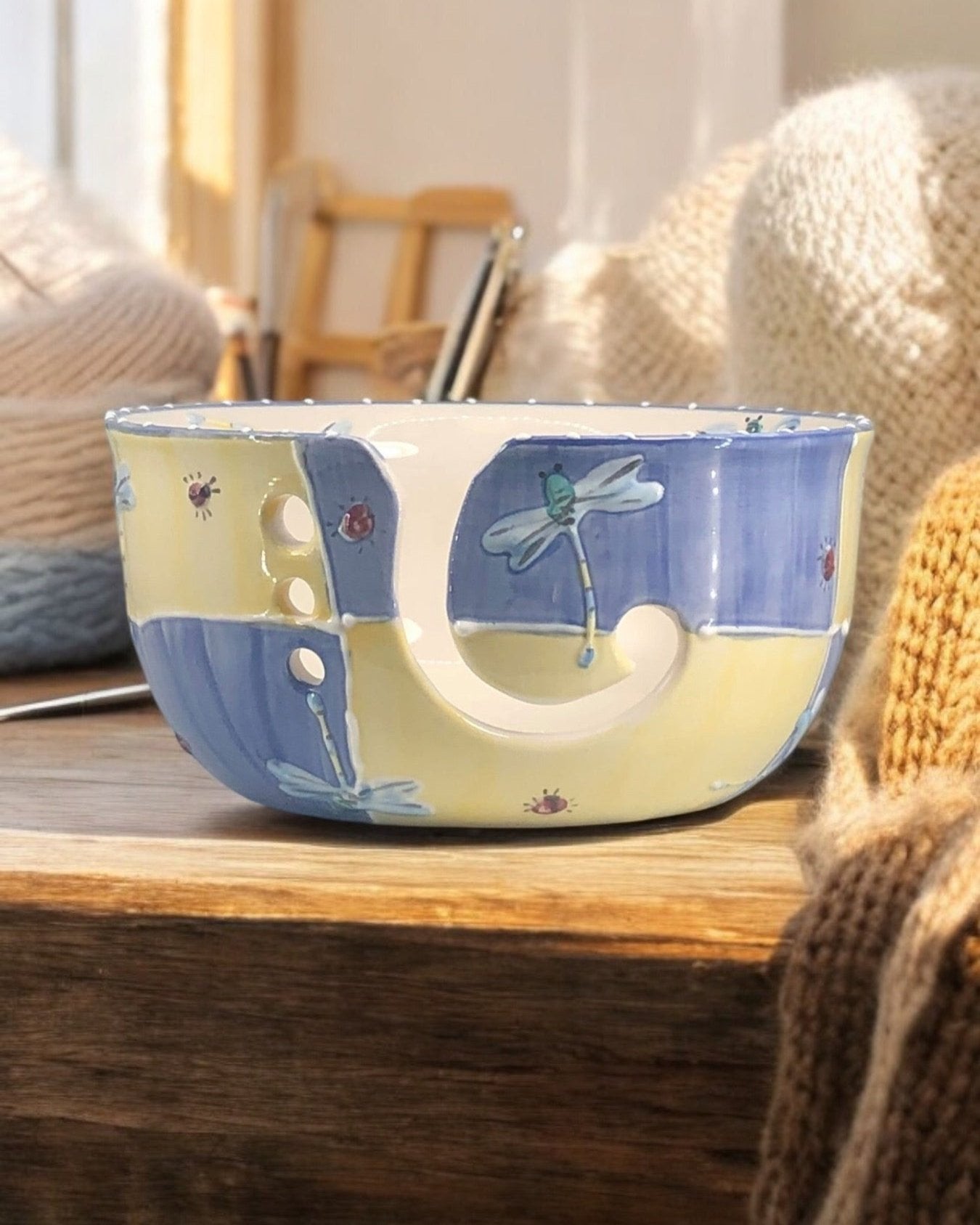 A hand-painted ceramic knitting bowl with dragonfly designs, containing yarn balls and knitting tools, placed on a wooden surface. Ladybird Ceramics Canada