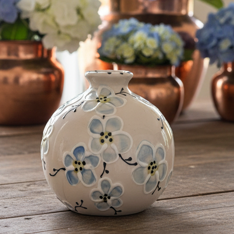 A small round ceramic vase with a white base, decorated with light blue and white floral motifs, sitting on a rustic wooden table with copper flower vases in the blurred background.