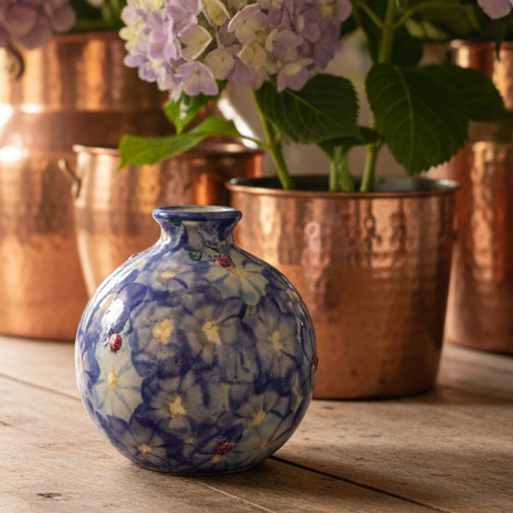 A small round ceramic vase in the foreground decorated with a blue floral pattern, set on a rustic wooden table with copper planters and purple hydrangeas in the background.