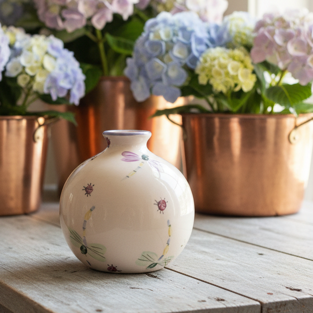 A small round ceramic vase with a cream-colored glaze and delicate hand-painted floral motifs sits on a rustic wooden table, styled in front of copper planters filled with pastel hydrangeas.