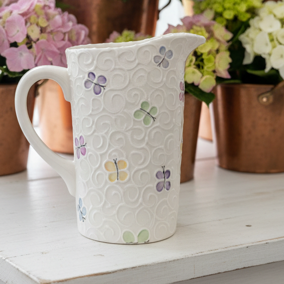 A white ceramic pitcher with embossed swirls and small pastel flower motifs, displayed on a white wooden surface with blurred pink, purple, and white hydrangeas in copper pots behind it.