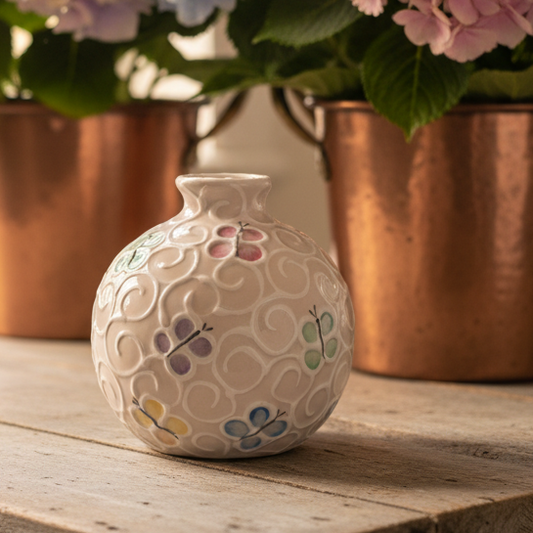 A small round ceramic vase with a textured bubble pattern and hand-painted pastel flowers, displayed on a rustic wooden table with copper planters and pink, blue, and purple hydrangeas in the background.