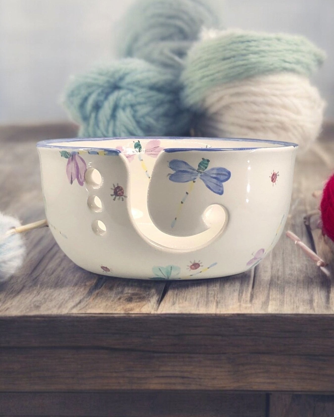 A hand-painted ceramic knitting bowl with blue dragonfly  painted design, containing yarn balls and knitting tools, placed on a wooden surface. Ladybird Ceramics Canada