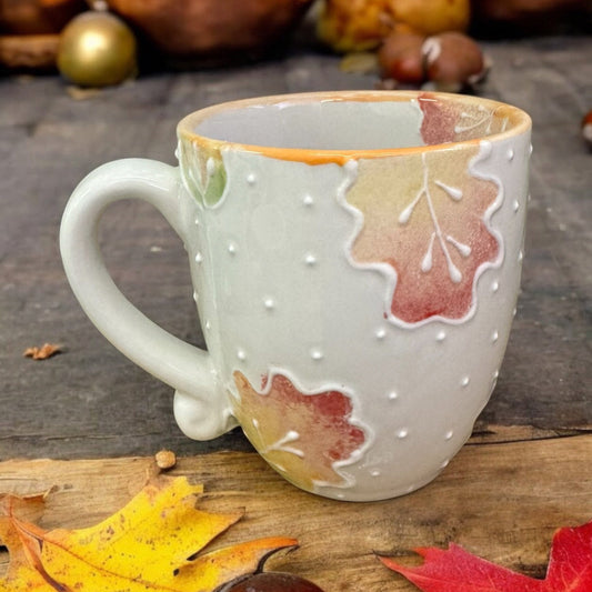 A small ceramic fall  mug with a hand-painted leaves design in shades of autumn, placed on a wooden surface surrounded by leaves and acorns. Ladybird Ceramics Original 