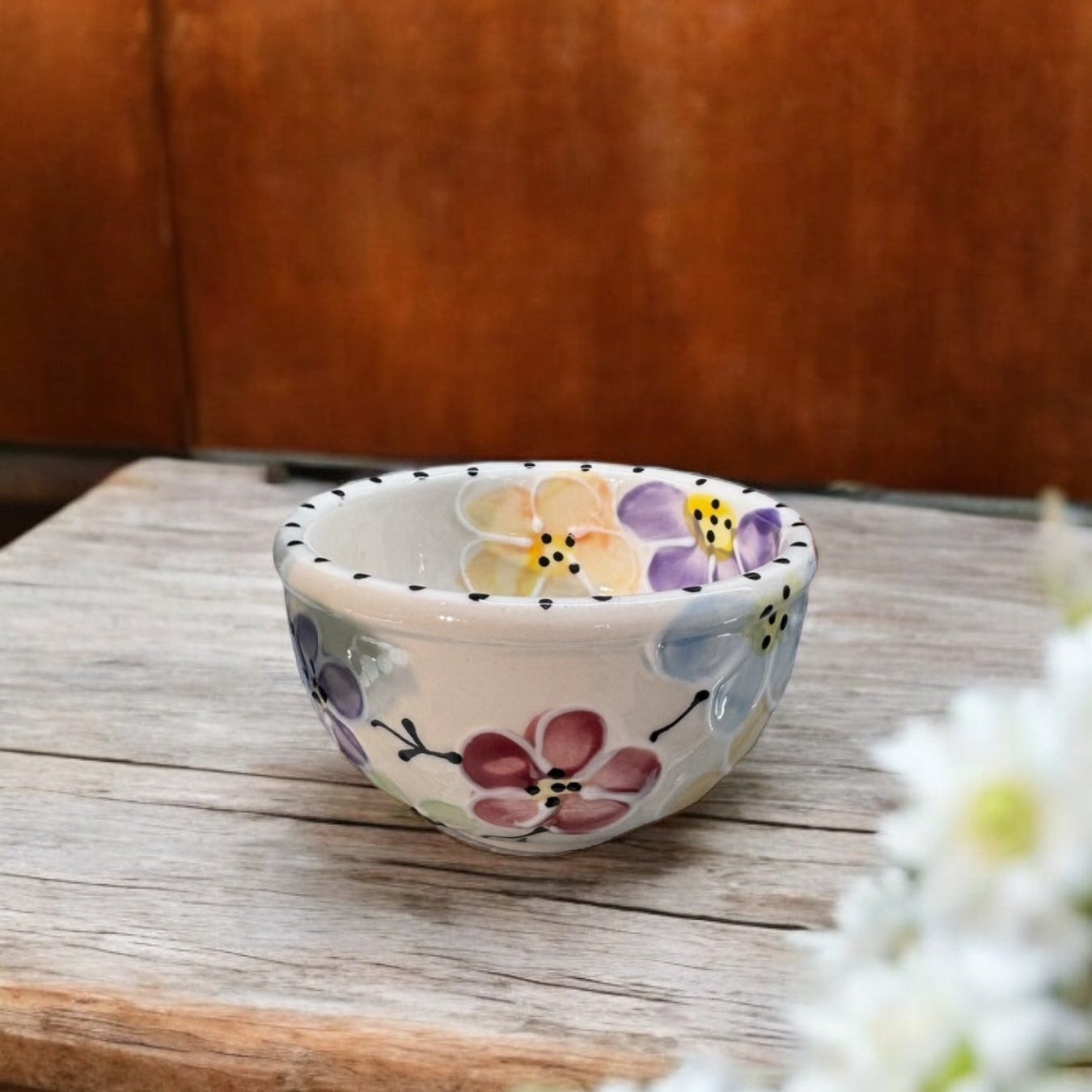A small ceramic bowl with a white base decorated with colorful hand-painted flowers in purple, pink, yellow, and blue, resting on a rustic wooden table.