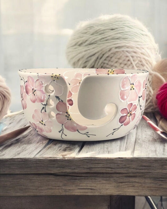 A hand-painted ceramic knitting bowl with pink cherry blossom floral designs, containing yarn balls and knitting tools, placed on a wooden surface. Ladybird Ceramics Canada