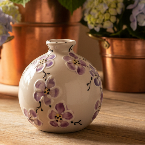 A small round ceramic vase with a narrow neck, decorated with hand-painted purple flowers and green leaves on a cream background, set on a wooden table with copper planters and hydrangeas in the background.