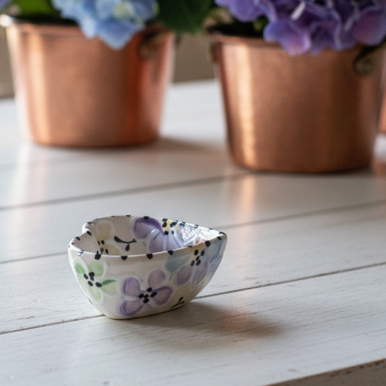 A small hand-painted ceramic bowl with a purple and green floral pattern sits on a white wooden table, with copper pots of blue and purple hydrangeas blurred in the background.