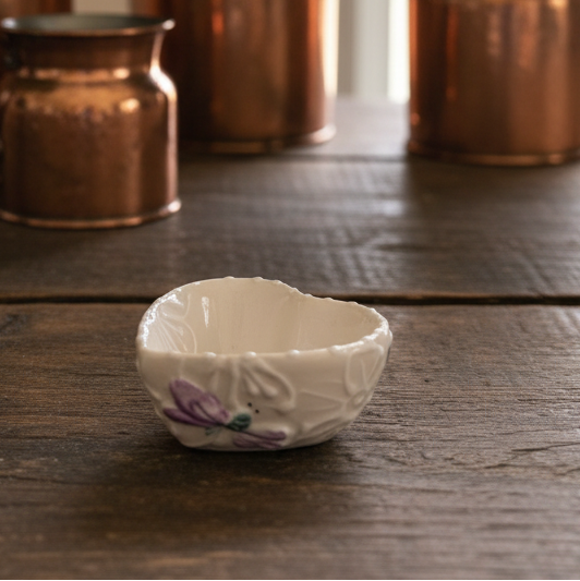 A small white ceramic bowl with a textured embossed floral pattern and a purple painted flower, set on a rustic dark wood table with copper pitchers and purple hydrangeas in the background.