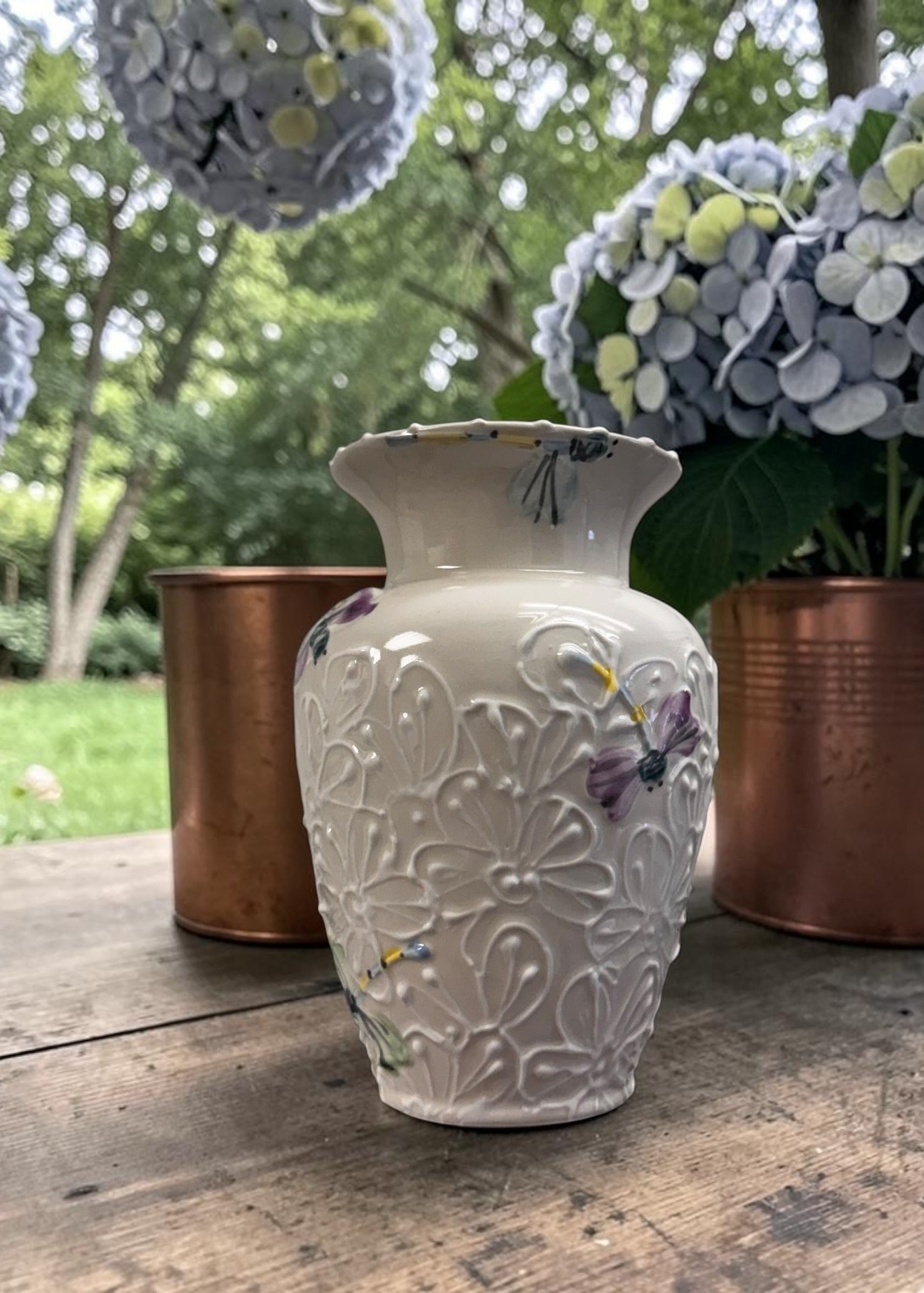 White decorative vase with dragonfly patterns on a wooden surface with potted plants in the background.