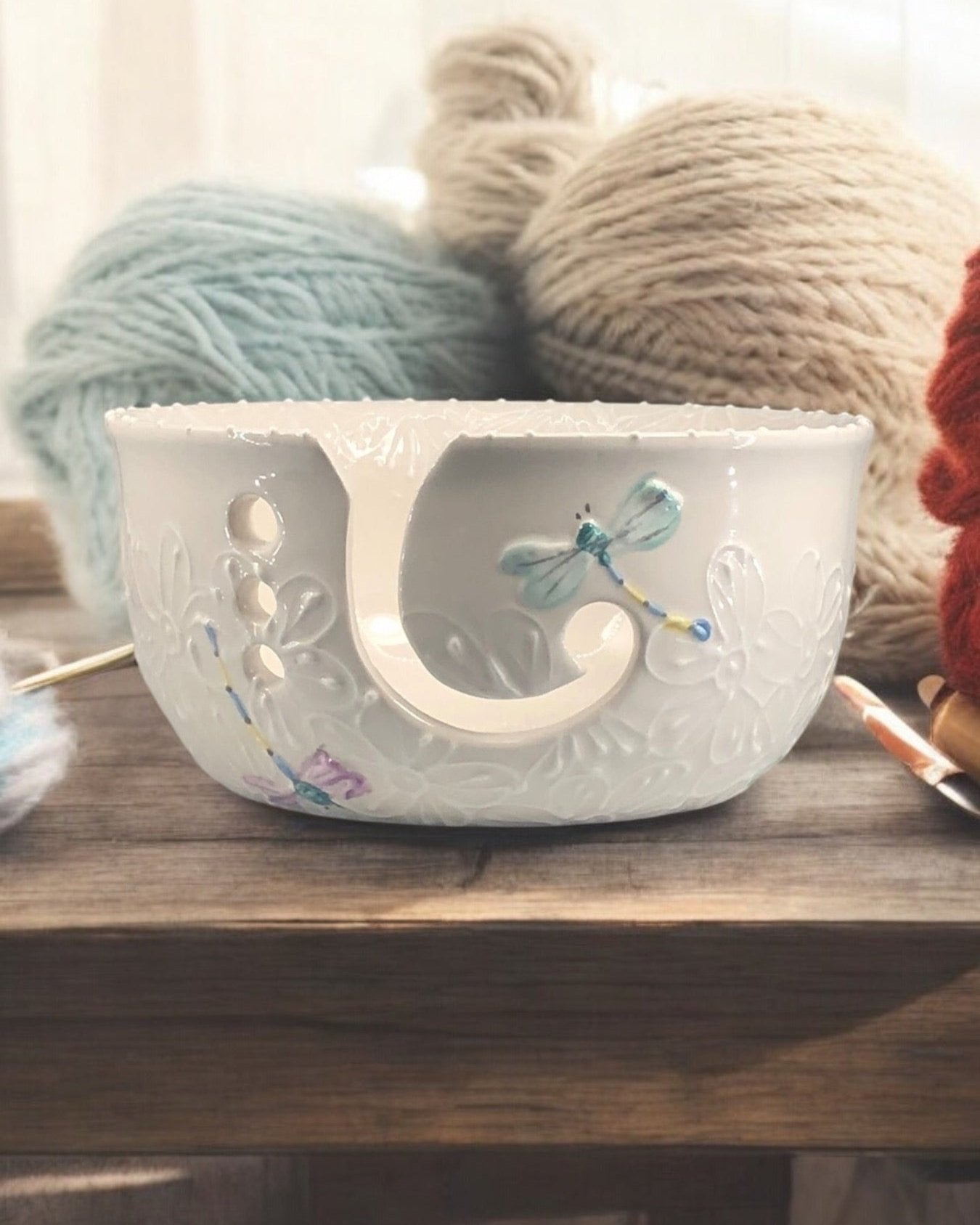 A hand-painted ceramic knitting bowl with dragonfly designs, containing yarn balls and knitting tools, placed on a wooden surface. Ladybird Ceramics Canada
